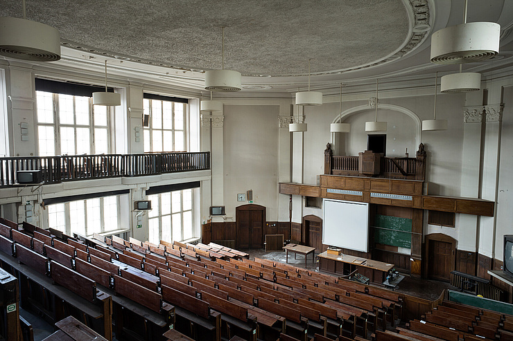 Das Bild zeigt einen großen, leeren Hörsaal in einem Gebäude mit historischem Flair. Der Raum ist mit Holzbänken und -tischen ausgestattet, die in Reihen angeordnet sind und zum Podium hinunterführen. An der Vorderseite des Raumes befindet sich ein Podium mit einem Pult und einer Tafel, auf der mathematische Formeln zu sehen sind. Über dem Podium hängt eine große Leinwand für Präsentationen.
