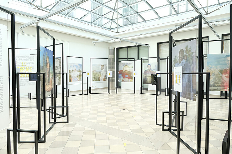 Das Bild zeigt eine Ausstellungshalle mit einem gläsernen Dach, das viel Tageslicht hereinlässt. Der Boden ist mit quadratischen Fliesen in einem Muster aus hellen und dunkleren Farben ausgelegt. In der Halle sind mehrere freistehende, schwarze Metallrahmen aufgestellt, die als Informationspods dienen. An diesen Rahmen sind Poster und Fotos befestigt, die verschiedene Personen und Szenen darstellen.