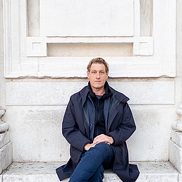 Joerg Fingerhut Joerg Fingerhut sitzt mit übergeschlagenen Bein vor einem Marmordenkmal und trägt eine dunkle Jacke.