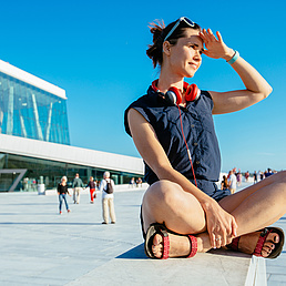 Young hipster woman traveler with red headphones resting on border after walking in Oslo city over Opera House urban background, happy, smiling, looking in sunset.